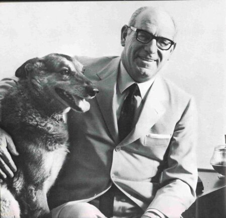 Uomo maturo in abito chiaro e cravatta, seduto accanto al suo cane di media taglia: sorride rivolto verso l’obiettivo mentre accarezza l’animale, in un ritratto in bianco e nero.