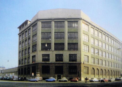 Grande edificio industriale in mattoni e cemento su più piani, con ampie finestre disposte simmetricamente; davanti al fabbricato, auto d’epoca parcheggiate e una strada urbana, sembra un capannone tessile degli anni ’60.