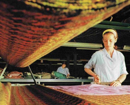 Operaia al lavoro in un laboratorio tessile mentre taglia con precisione un tessuto colorato, circondata da rotoli di stoffa e postazioni di lavoro