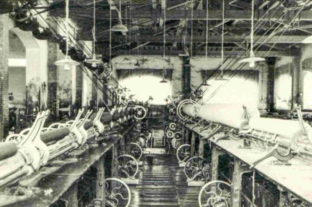 Interno di una fabbrica tessile italiana del primo Novecento con telai meccanici e cinghie di trasmissione sospese, tipica dell’industria laniera o cotoniera dell’epoca.